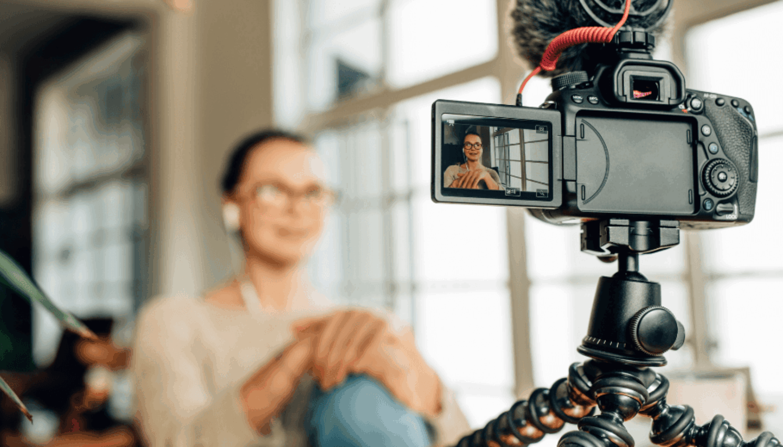 A DSLR camera on a flexible tripod in sharp focus in the foreground, with its flip-out LCD screen showing a woman recording herself. In the background, a blurred woman with glasses and white earbuds sits smiling in a bright, modern room.